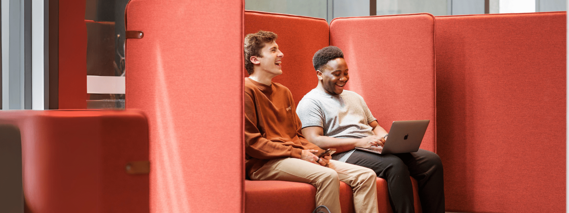 Two male students are sitting together on a bench seat. One student is using a laptop.