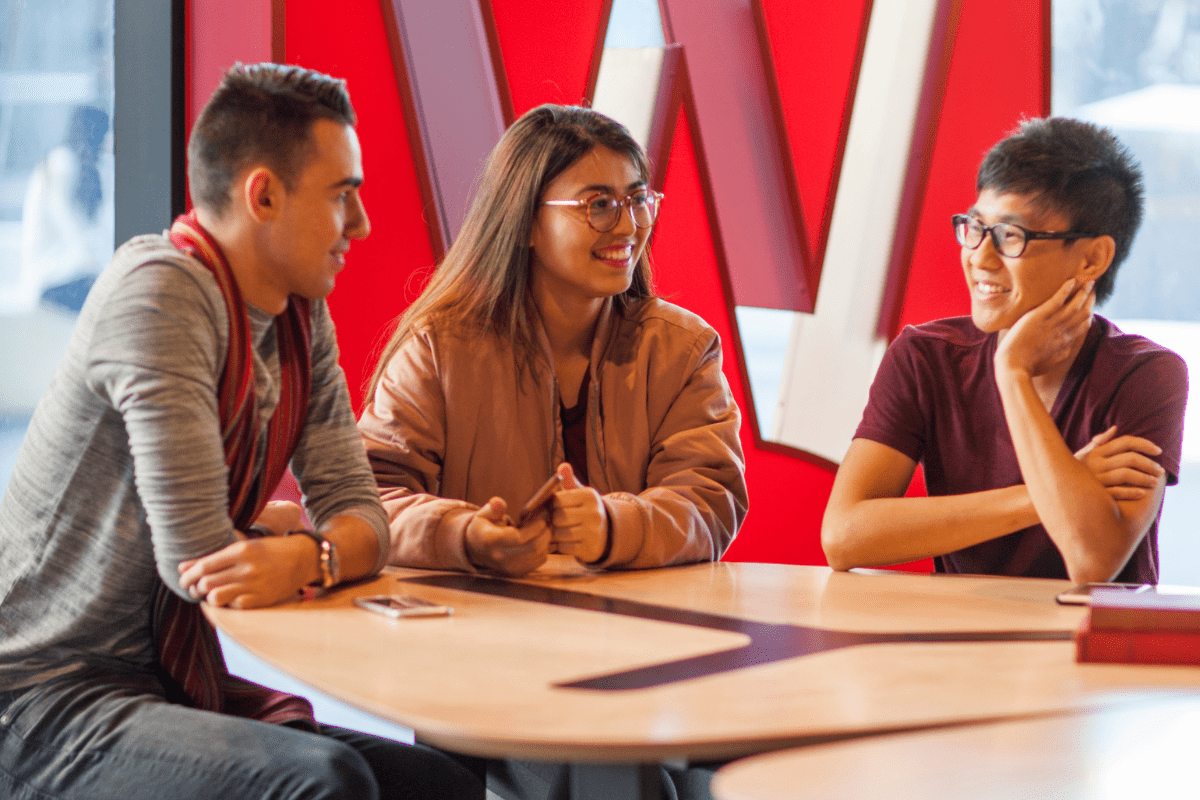 Three students are sitting together chatting at a desk.