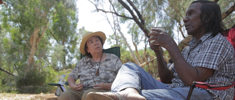 A woman (Alexis Wright) and a man (Clarence Walden) are seated next to each other in bushland