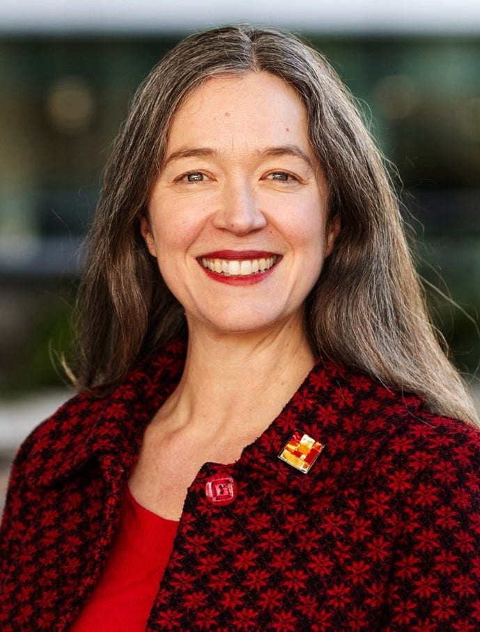 Kate wears a square necked black top, with a colourful necklace. Her long hair is swept back over her shoulders. She smiles at the camera.