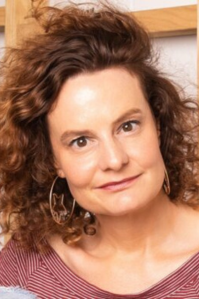 Felicity has medium length brown wavy hair. She wear a striped red and white shirt and hoop earrings.