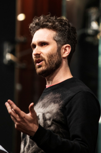 Ben wears a black sweater. He has curly brown hair and a moustache and beard. He is looking off-camera, speaking to an audience.