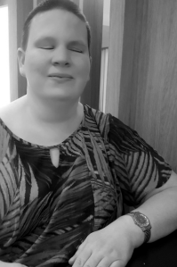 Amanda Tink wears a loose blouse with leaves printed on it. They have a shortcut buzz haircut and faces toward the camera, with a slight smile.